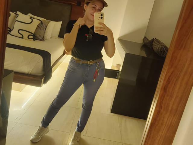 MabyLegrand with a cap, black shirt, blue jeans, and white sneakers, holding a phone, showing a mirror selfie with a belt and keys in the room.