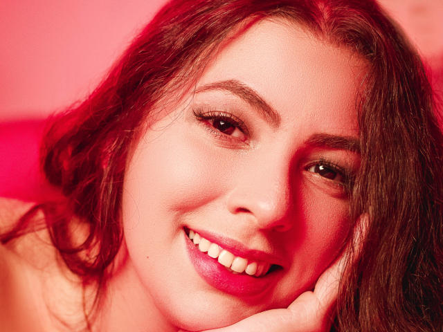 VanessaClose's face, red lit, with wavy brown hair, pink lipstick, smiling, showing white teeth, hand resting on cheek, brown eyes, subtle makeup, soft skin, slightly flushed.