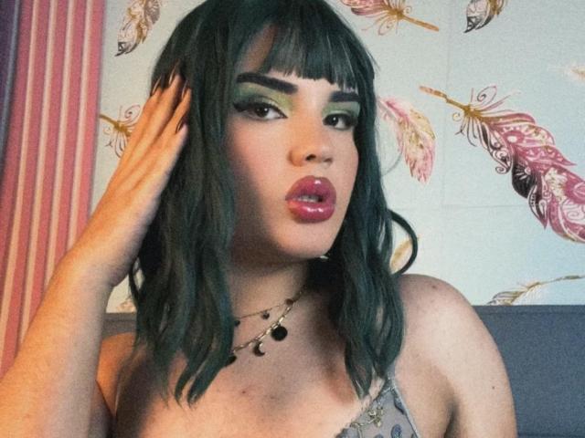 GlossyyBratz mit dunkelgrünen Haaren, auffälligem grünen Lidschatten, pinkem Lippenstift und einer Hand im Haar, trägt ein graues Oberteil und eine Halskette und zeigt ihre Brüste..