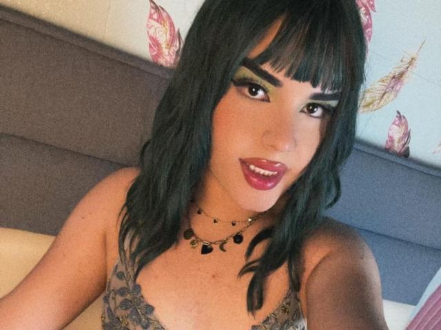 A GlossyyBratz tem cabelo preto ondulado, delineador marcante e batom vermelho. Ela está vestindo uma blusa de renda com um colar preto, mostrando seus seios. Ela está sorrindo para a câmera..