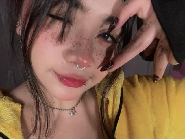 AmaraKazumi with freckles, nose ring, and lip piercing, winking, wearing yellow hoodie, long dark nails, pink lipstick, necklace, and black hair. Her face is slightly flushed.
