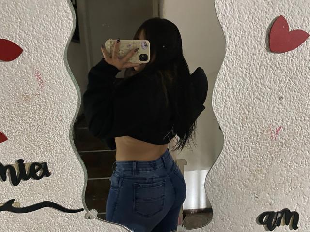AmaraKazumi with long dark hair, wearing a black cropped hoodie and blue high-waisted jeans, taking a mirror selfie showing her bare back and butt, holding a phone.