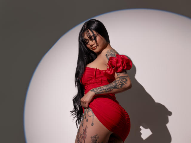 LenaLegrand, con lunghi capelli neri, vestito rosso con una spalla scoperta, tatuaggi sul braccio e sulla coscia, in piedi, con un'ombra proiettata su un muro bianco, che guarda verso la fotocamera, con una mano sul vestito, illuminata dalla luce del sole..