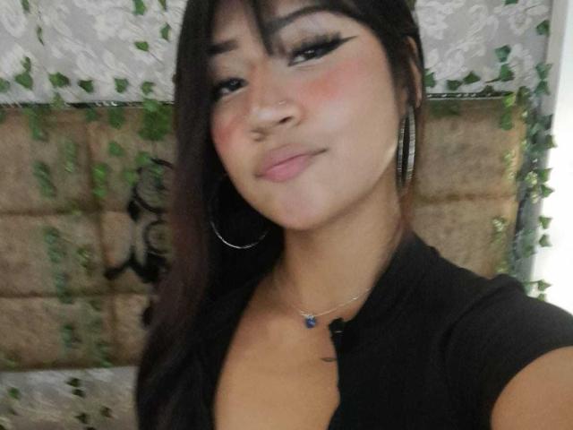 BonnieBond with dark hair, thick eyeliner, pink blush, hoop earrings, black top, and a blue necklace, smiling slightly, showing her cute ass in the selfie.