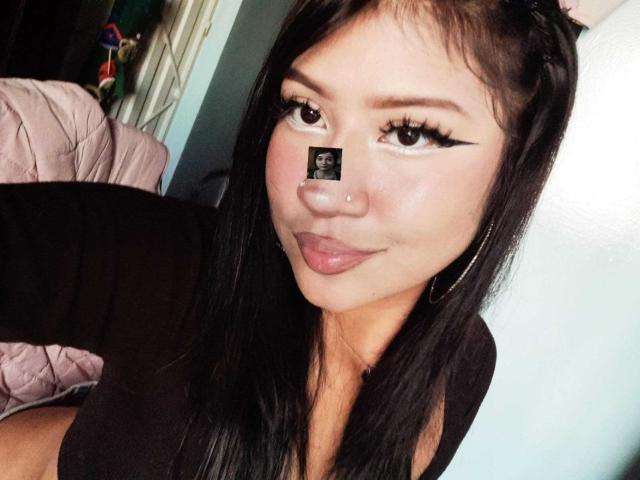 BonnieBond heeft zwart haar, lange wimpers en een neus met een klein stickertje. Ze draagt een zwarte top, met roze lakens op de achtergrond..