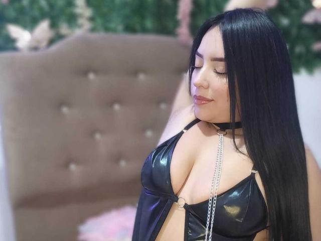 AdaraCute69, avec de longs cheveux noirs, portant un soutien-gorge en cuir noir brillant, des chaînes et un collier noir, montrant ses grandes seins, souriant les yeux fermés..