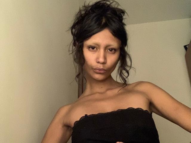 AliceFirek has a slim, topless physique with small tits, dark messy hair, and a serious expression, wearing a black strap with lace. Her brown eyes are focused.