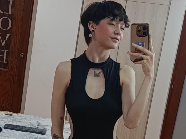 LissyLuxe with short black hair, small tattoos, and pink cheek, wearing a sleeveless black top with a keyhole, taking a mirror selfie with a phone, showing her tits.