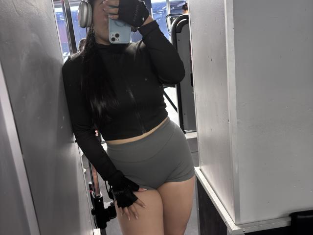 MiaDuboisX, con cabello largo y negro, vestida con una camiseta corta de manga larga negra y pantalones cortos grises de cintura alta, sosteniendo un teléfono en un selfie frente al espejo, mostrando su trasero..