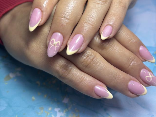 De handen van MiaDuboisX, met glanzende roze en gele nagellak, met witte strikjes op sommige nagels, rusten op een blauw oppervlak. Haar vingers zijn licht glanzend en ze draagt een rode polsband..