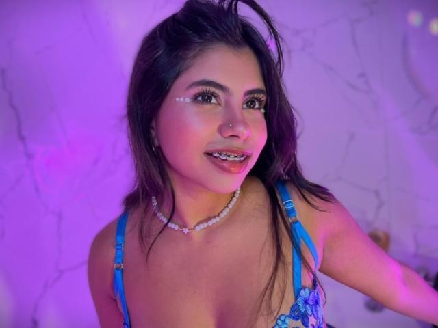 CelesteMorel with dark hair, blue lingerie, pearl necklace, face paint dots, nose ring, and purple lighting, showing her tits and smiling.
