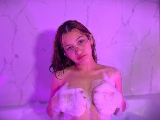 Elodie Gardner, con i capelli castani bagnati, che indossa gli occhiali, tiene delle bolle saponose sopra il seno in una vasca da bagno illuminata di viola. I suoi occhi sono rivolti in avanti, le labbra leggermente dischiuse..