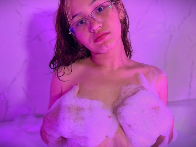 Elodie Gardner, con i capelli castani bagnati, che indossa gli occhiali, tiene tra le mani dei seni pieni di sapone, illuminata da una luce viola, guarda verso la telecamera, con le labbra rosa leggermente dischiuse, in una vasca da bagno..