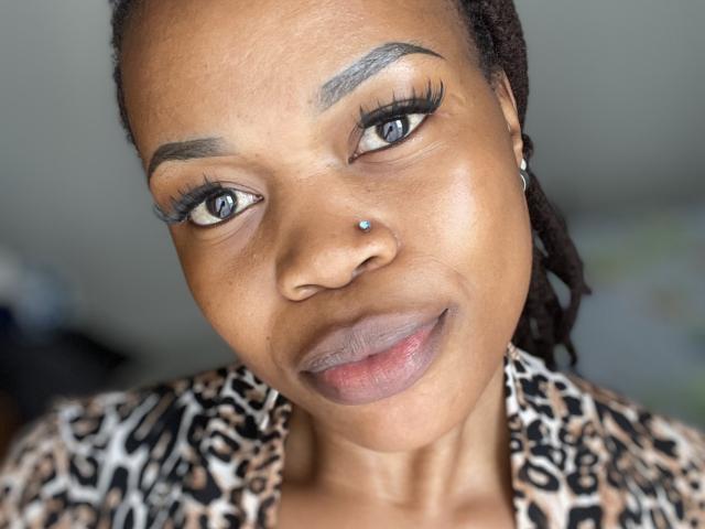 Sashie's face, dark brown skin, blue eyes, thick black eyebrows, long eyelashes, nose piercing, leopard print shirt, smiling slightly, black dreadlocks, shiny lips, subtle makeup.