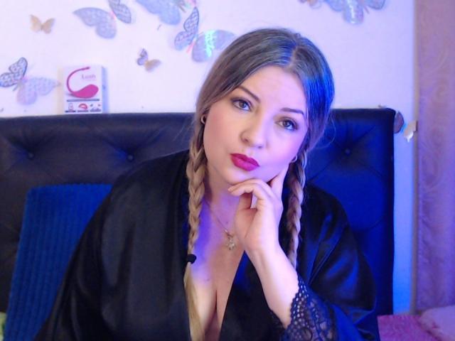 NatalyHami with blonde braids, wearing a black silk robe, blue eyes, pink lipstick, hand on chin, black lace sleeve, sitting on a blue bed, looking at camera.