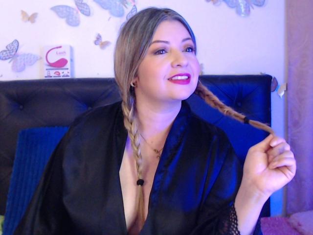 NatalyHami con i capelli intrecciati, indossa un abito di seta nera, mostra il décolleté, tiene una treccia, sorride con rossetto rosa, ha gli occhi azzurri e la pelle chiara..