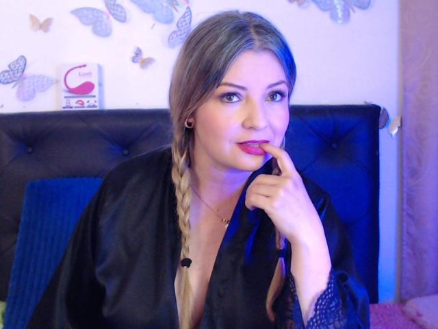 NatalyHami, avec des tresses blondes, une robe en satin noir, des yeux bleus, mordant sa lèvre, regardant la caméra, avec une poitrine légèrement visible, un tête de lit noir capitonné, des papillons sur le mur, un éclairage bleu subtil..