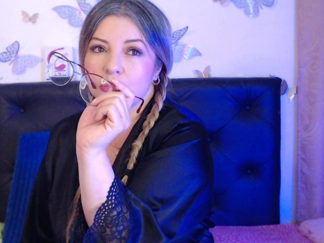 NatalyHami, avec des cheveux tressés, portant une robe en soie noire, tenant des lunettes près de ses lèvres, un tête de lit bleu avec des motifs, regard séduisant, yeux bleus, boucles d'oreilles cercles..