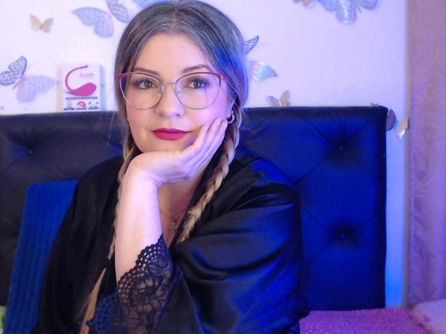 NatalyHami with braided blonde hair, red lipstick, glasses, black lace sleeves, and a black satin robe, resting her chin on her hand, looking cute and sexy.
