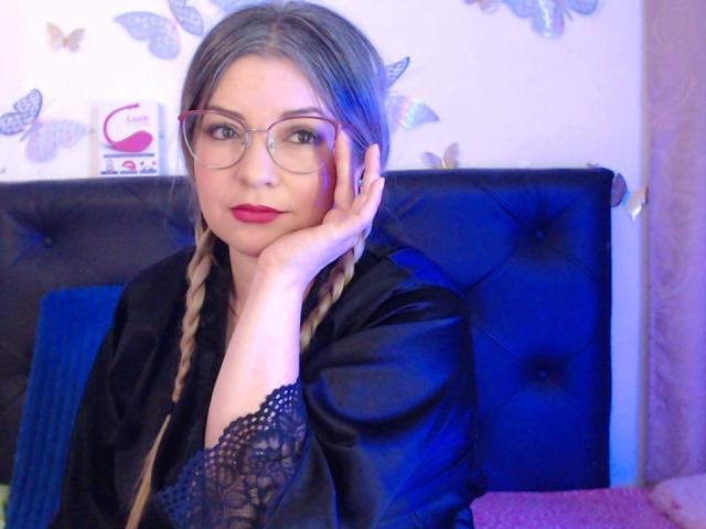 NatalyHami avec des tresses, des lunettes, du rouge à lèvres rouge, une chemise en dentelle noire, une main sur la joue, une tête de lit bleue, et des papillons sur le mur, ayant l'air mignonne et sexy..