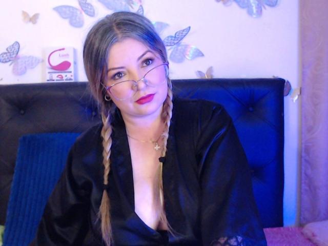 NatalyHami with glasses, braided blonde hair, black satin robe, cleavage, red lipstick, black tufted headboard, blue pillow, butterfly wall decals, necklace, soft blue lighting.