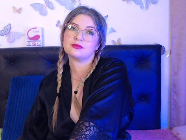 NatalyHami with braids, glasses, red lipstick, black lace sleeves, black satin robe, blue headboard, butterfly wall decor, and gold earrings, sitting on bed, smiling slightly.