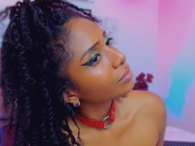 MeganWilliams with dark curly hair, green eyeshadow, nose ring, red collar, and earrings, looking over shoulder with a subtle smile, showing bare shoulders.