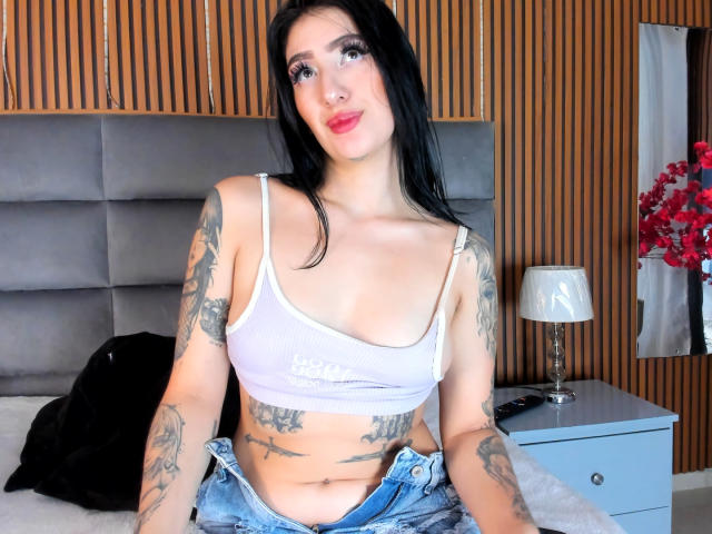 TifanyDiaz with black hair, white crop top, tattoos, and blue jeans, showing her tits and belly button, sitting on a bed with a lamp and red flowers.