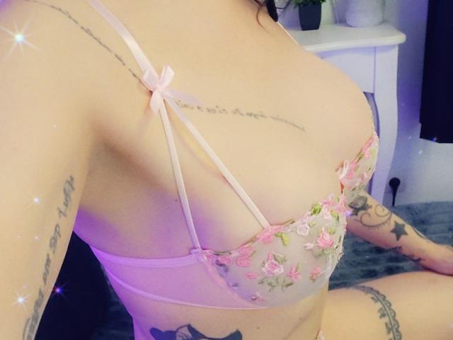ElixirShanna's tits in a pink floral bra, showing tattoos on her arm and side, with a pink bow on the strap. Her skin is light, and she's sitting.