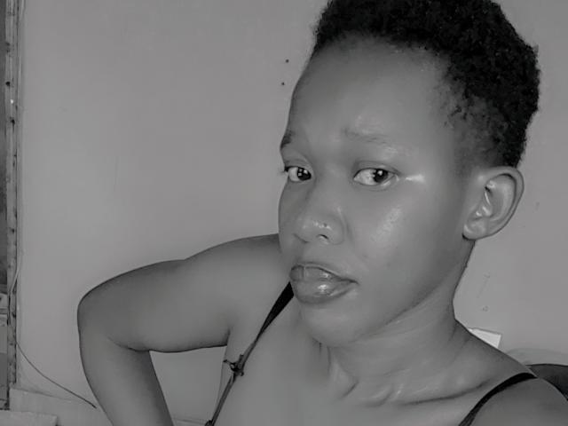 Black-and-white photo of DivineDivvah with short curly hair, glossy lips, and visible shoulder strap, looking slightly to the side with a neutral expression. Her tits are small.