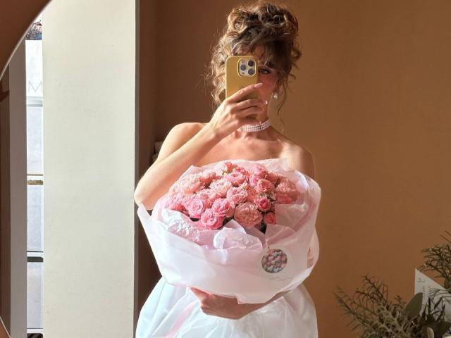 PortiaKomer, con cabello castaño rizado, sosteniendo rosas rosadas con un vestido blanco, tomando una selfie en el espejo, cubriendo sus pechos con el teléfono, un collar rosa, una pared beige, una ventana y una planta en el fondo..