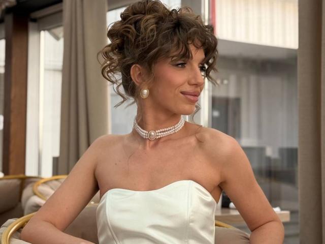 PortiaKomer hat lockiges, braunes Haar, Perlenohrringe und eine Perlenkette. Sie trägt ein weißes, trägerloses Kleid mit kleinen Brüsten, lächelt sanft und hat helle Haut..