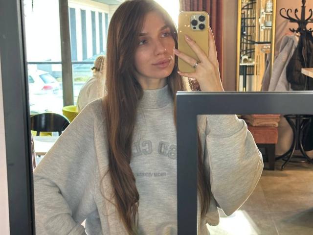 PortiaKomer with long brown hair, wearing a grey sweatshirt, holding a gold iPhone in front of a mirror, showing her face and slightly puffy ass in the reflection.