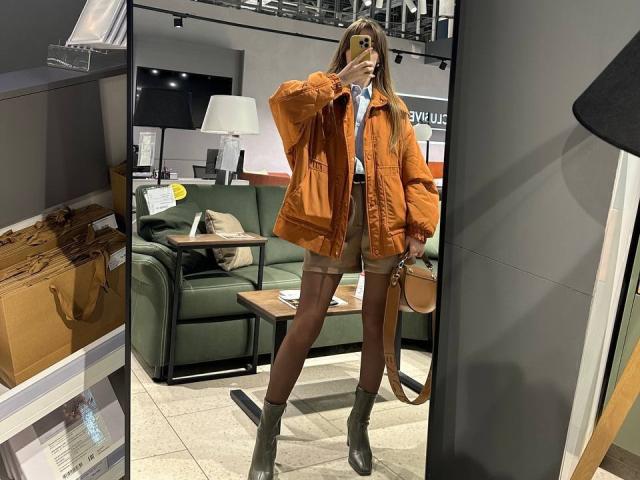 PortiaKomer stands in a store, wearing an orange jacket, brown shorts, green boots, and a white shirt, holding a phone, with a brown handbag on her right.