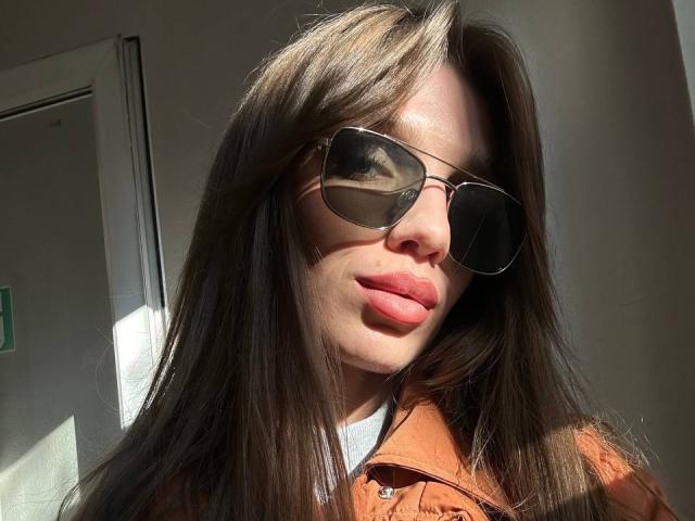 PortiaKomer, con lunghi capelli castani, indossa occhiali da sole da aviatore e una giacca marrone. La luce del sole illumina le sue labbra rosa e la sua camicia bianca. Le sue natiche non sono visibili..