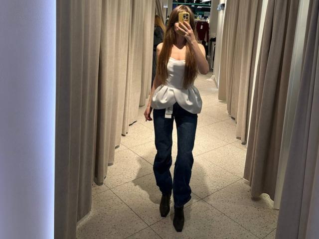 PortiaKomer, com cabelos longos e ruivos, vestindo uma blusa branca sem alças com mangas bufantes, jeans azuis e botas pretas, tirando uma selfie no espelho em uma loja de roupas..