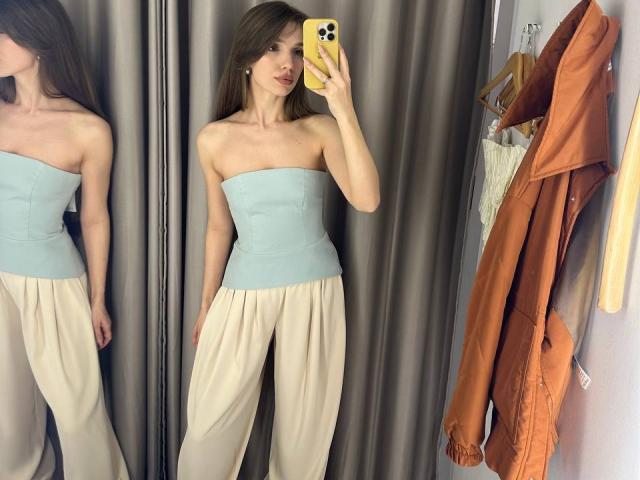 PortiaKomer has small tits, wearing a strapless light blue top and loose beige pants, taking a mirror selfie with a yellow phone, showing an orange coat hanging nearby.