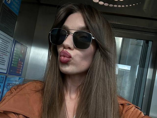 PortiaKomer mit langen, braunen Haaren, die dunkle Sonnenbrillen und eine braune Jacke trägt, zieht ihre Lippen zu einem schmollenden Kuss zusammen. Ihre Brüste sind leicht unter der Jacke sichtbar..