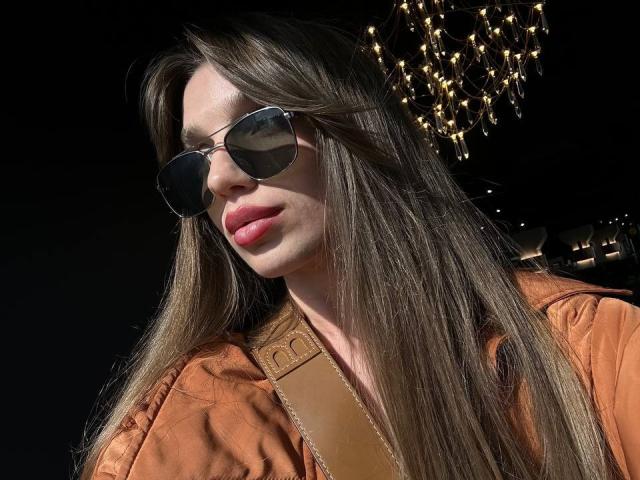 PortiaKomer with long brown hair, wearing round sunglasses, orange jacket, and pink lipstick, has a tan shoulder strap, and a string of lights in the background.