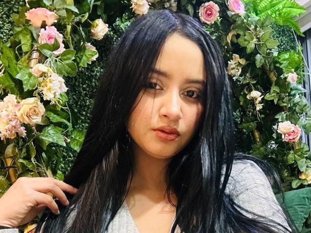 GraceBlue69 with long black hair, pouty lips, light skin, wearing a gray top, surrounded by pink roses and green leaves, looking slightly to the side with a hand in hair.