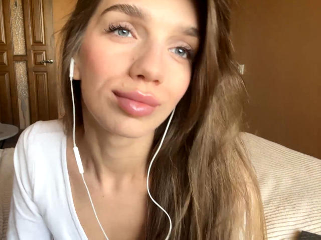 PortiaKomer tiene ojos azules, cabello largo castaño, audífonos blancos y labios rosados, viste una blusa blanca, sonríe ligeramente y tiene un ligero rubor en las mejillas..