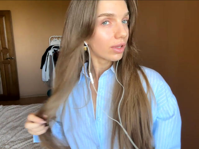 PortiaKomer with long brown hair, blue eyes, and pink lips, wearing a blue shirt, holding hair, white earbuds in, looking to the side, in a softly lit room.