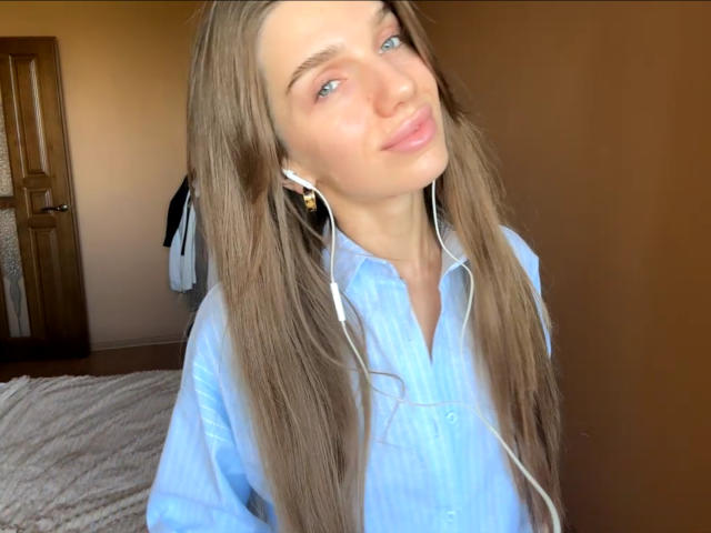 PortiaKomer, avec de longs cheveux bruns, des yeux bleus et des lèvres roses, portant une chemise bleu clair, des écouteurs blancs et affichant un léger sourire, dans une pièce aux murs bruns..