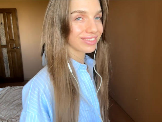PortiaKomer tiene el pelo largo y castaño, ojos azules y una sonrisa encantadora. Lleva una camisa azul con auriculares blancos. Sus senos no son visibles. Está de pie en una habitación..