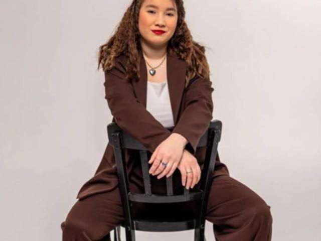 Dawne Norsworthy, con cabello castaño rizado, lápiz labial rojo, traje marrón, camisa blanca, sentada en una silla negra, con las manos sobre el asiento, sonriendo, y llevando un collar, un anillo y dos pulseras..