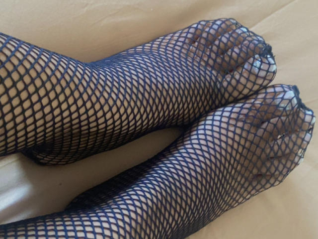 Os pés da EloCherie, usando meias-calças de rede preta, mostrando seus dedos e unhas através do tecido estampado, estão sobre uma superfície bege..