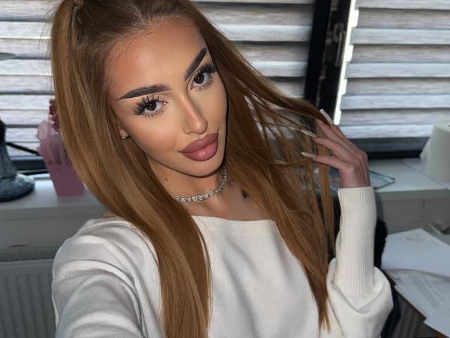 MarissaFox mit langem, glattem, braunem Haar, die einen weißen Pullover, eine Diamantkette und einen Eyeliner trägt, hält sich ihr Haar zur Seite, um ihre vollen, rosafarbenen Lippen und ihr hübsches Hinterteil zu zeigen..