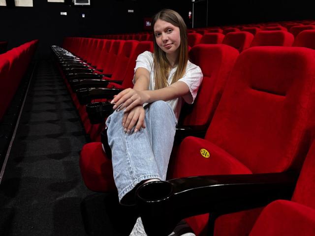 BabeLumi with long brown hair, white t-shirt, and blue jeans, sitting in a red cinema seat, legs crossed, black armrests, black carpet, and a slight smile.
