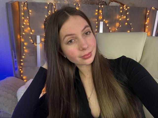 BabeLumi with long brown hair, black top, and subtle smile, showing her tits in a selfie with warm fairy lights in the background. She has light skin and pink lips.