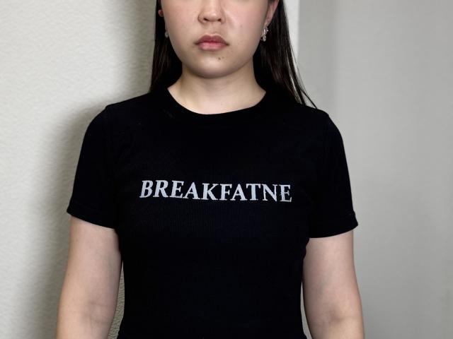 MariLerew has a black shirt with "BREAKFAST" in white letters, standing with long brown hair, pale skin, and subtle makeup, wearing small earrings.